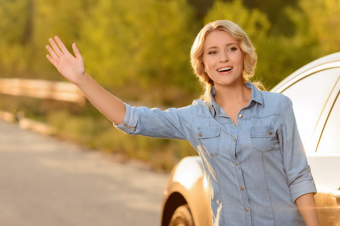 A woman waving to someone while standing next to her car.